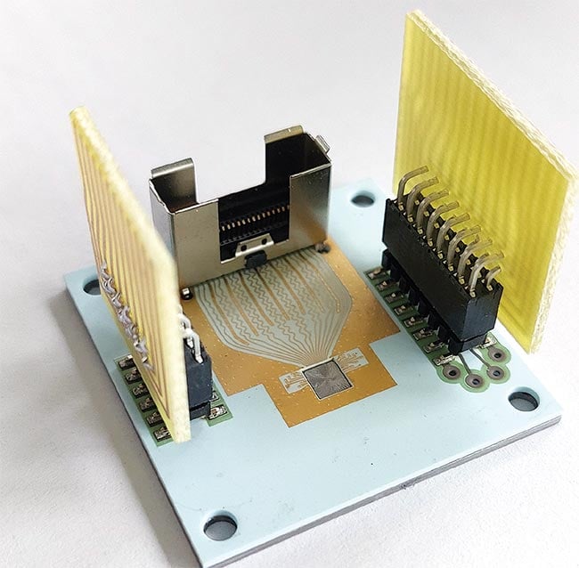 Figure 2. A ceramic printed circuit board (PCB) is bonded to a molybdenum heatsink of a similar size. The PCB carries the VCSEL and driver chip (center). The wings are plugs for thermal management components. The plug in the back will connect to the gigahertz driver signal. See Reference 4. Courtesy of TU Ilmenau.