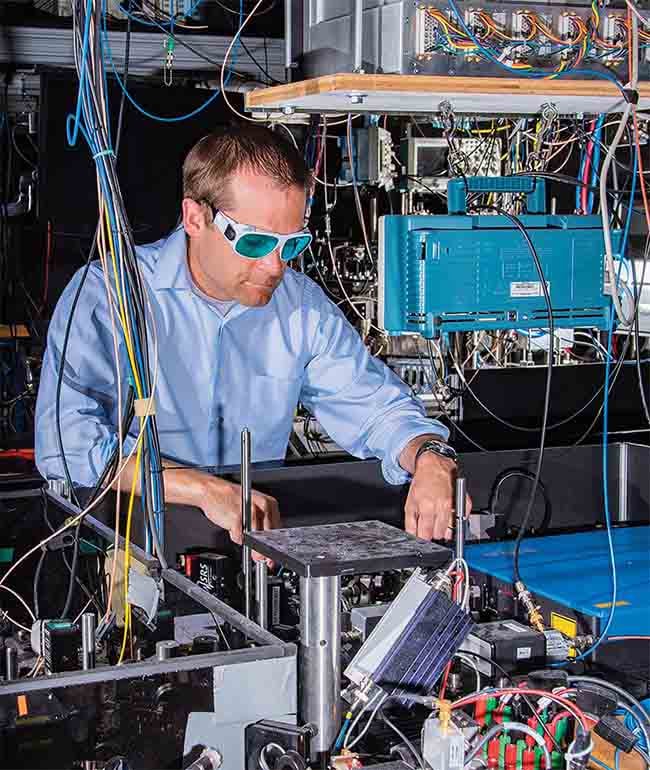National Institute of Standards and Technology (NIST) physicist Andrew Ludlow and colleagues achieved atomic clock performance records in a comparison of two ytterbium optical lattice clocks. The photo shows the laser systems used in both clocks (foreground) and the main apparatus for one of the clocks (behind). Courtesy of NIST/Burrus via TOPTICA Photonics AG.
