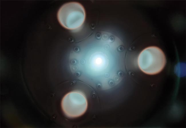 An electrodeless Z-Pinch discharge plasma source. The bright spot is the 13.5-nm emitting ‘pinch.’ Three plasma ‘return loops’ emit brightly in the visible region (below). Courtesy of Energetiq.