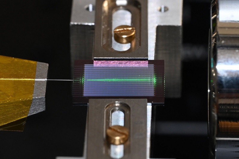 The experimental setup shown here incorporates on-chip optical parametric oscillator (OPO) technology to generate a frequency comb of laser-like light covering a wide range of frequencies with very little input energy. In this image, the chip includes about 20 OPOs, and one of them is being tested. An optical fiber is shown to the left of the chip and a free-space objective to the right. Courtesy of Caltech/Alireza Marandi.